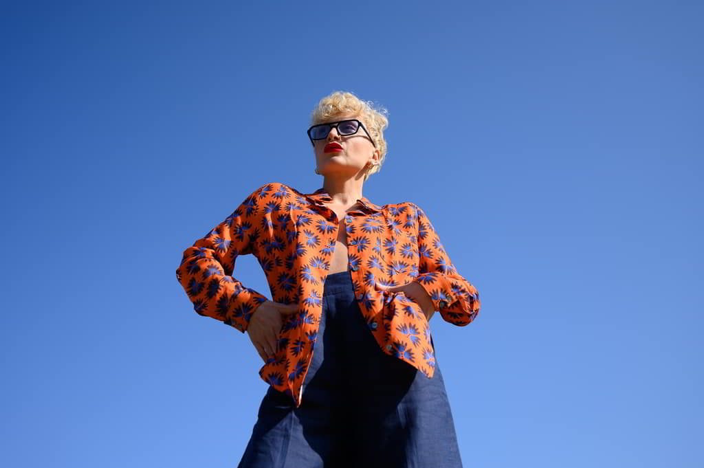 Chemise Chemise à motifs floraux en viscose EcoVero orange, bleu, motifs floraux en viscose EcoVero certifié EcoVero avec lunettes de soleil noires, jupe bleue foncée porté par femme, cheveux stylés debout, mains sur les hanches — 3/4 — extérieur, ciel bleu clair, ambiance décontractée plein air