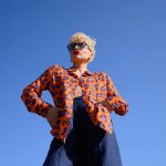 Chemise Chemise à motifs floraux en viscose EcoVero orange, bleu, motifs floraux en viscose EcoVero certifié EcoVero avec lunettes de soleil noires, jupe bleue foncée porté par femme, cheveux stylés debout, mains sur les hanches — 3/4 — extérieur, ciel bleu clair, ambiance décontractée plein air